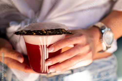 A person holding a red cocktail garnished with a thin chocolate strip topped with crushed nuts.