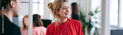 A woman wearing a red blouse is talking to a colleague in a modern, brightly lit office for teamwork and corporate messaging