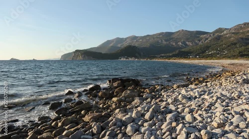 A pebble shoreline, gentle waves lapping against the stones, a tree-covered headland in the distance, and the afternoon sun.