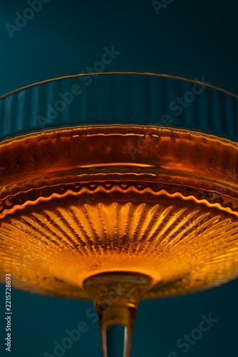 Extreme close-up macro of an amber cocktail in a textured, vintage crystal coupe glass with fine ribbed patterns and warm glowing light against a dark teal background.