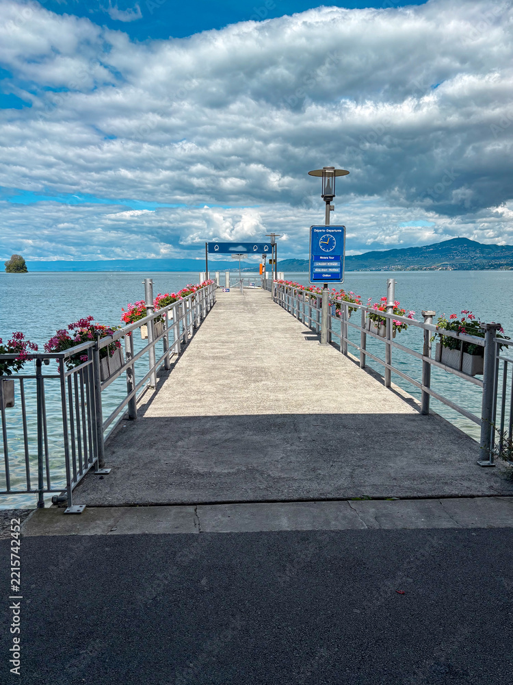 Fototapeta premium Lake geneva pier extending into calm waters under a dramatic cloudy sky, with blooming flowers lining the railings and distant mountains creating a serene travel destination scene