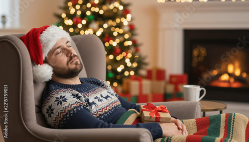 Tired Man in Santa Hat Sleeping in Armchair by Christmas Tree and Fireplace