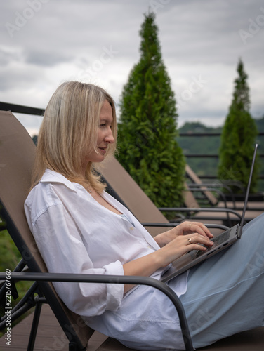 Woman relaxing on a deck chair, typing on her laptop, embracing remote work and digital nomad lifestyle with a panoramic outdoor view of mountains and nature. Vertical photo