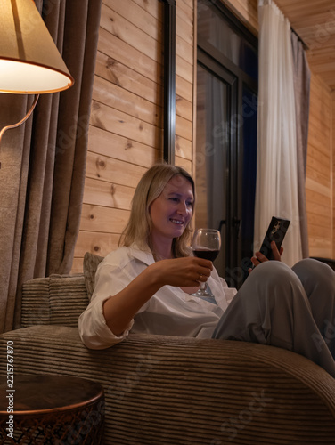 Smiling woman sitting comfortably on a sofa at home during the evening, holding a glass of red wine and using a smartphone for relaxation and entertainment. Vertical photo