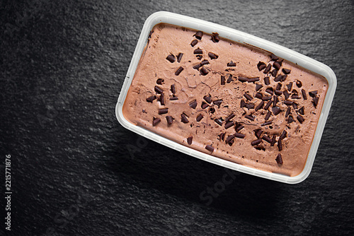 Chocolate ice cream with chocolate chips in a plastic container, photographed from above on a dark textured background.