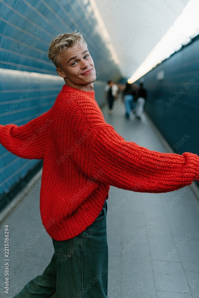 Fototapeta premium Blonde young man wearing a red oversized knit sweater spinning in a subway tunnel for fashion background and poster