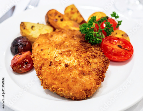 Crispy fried cutlet with roasted tomatoes, potatoes, and herbs on a white plate. AI