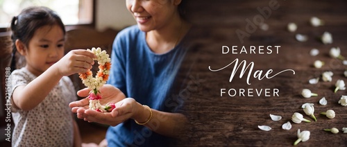 Thai daughter giving jasmine garland to mother for Mothers Day celebration, traditional Phuang Malai expressing deep respect, family bonding and love with dearest Mae text over a rustic wooden table.
