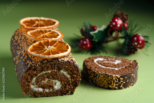 Christmas yule log cake with cream filling, dried oranges, and a cut slice, styled with festive holiday decorations on a green background. Warm holiday feeling