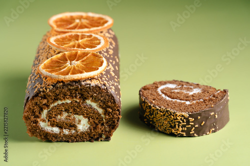 Homemade chocolate cream roll cake topped with dried oranges and a single cut slice on a light green background. Smooth lighting and horizontal layout