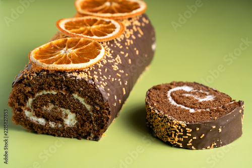Chocolate sponge cake roll with cream filling and a cut slice, garnished with dried citrus. Styled at a 45-degree angle on a textured green fabric background
