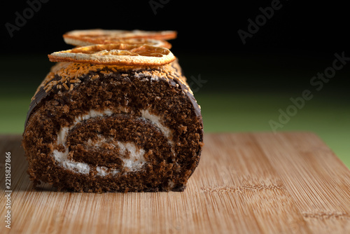Delicious chocolate swiss roll cake with cream filling and dried orange slices on a wooden board. Close up food photography with green background