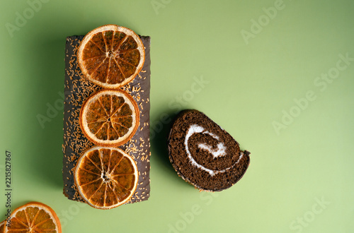 Top view of a chocolate swiss roll cake decorated with dried oranges and a cut slice beside it. Flat lay styling on a vibrant green background with copy space