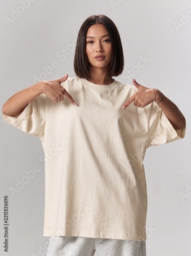 Woman wearing an oversized white t-shirt while pointing towards her chest against a plain studio background.