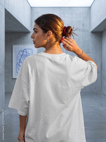 Rear view of a young woman wearing a white oversized t-shirt standing in a minimalist concrete art gallery space.