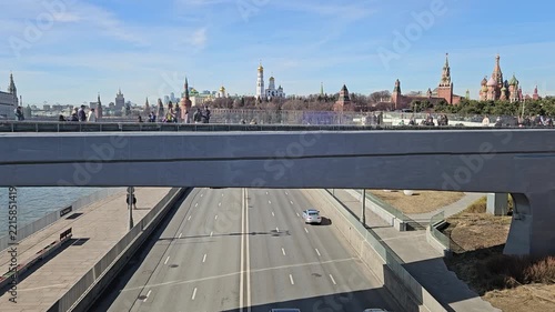 MOSCOW - MAR 22, 2026: Clear sky above busy highway over Moscow River. Cars move beneath concrete fly bridge. Red towers stand tall in distance. City skyline visible behind waterway near Kremlin.