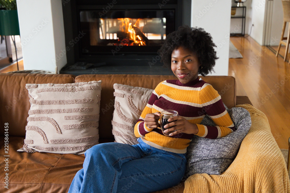 Naklejka premium Female sitting on leather couch by fireplace holding glass mug wearing striped sweater and jeans