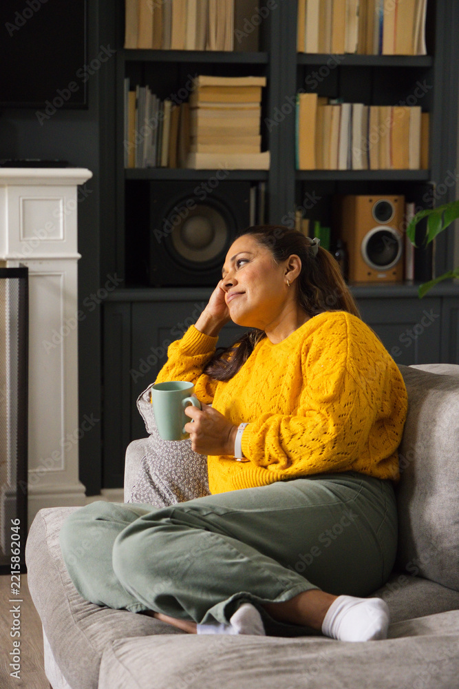 Fototapeta premium African woman wearing mustard sweater sitting on gray sofa holding light-green mug, gazing right