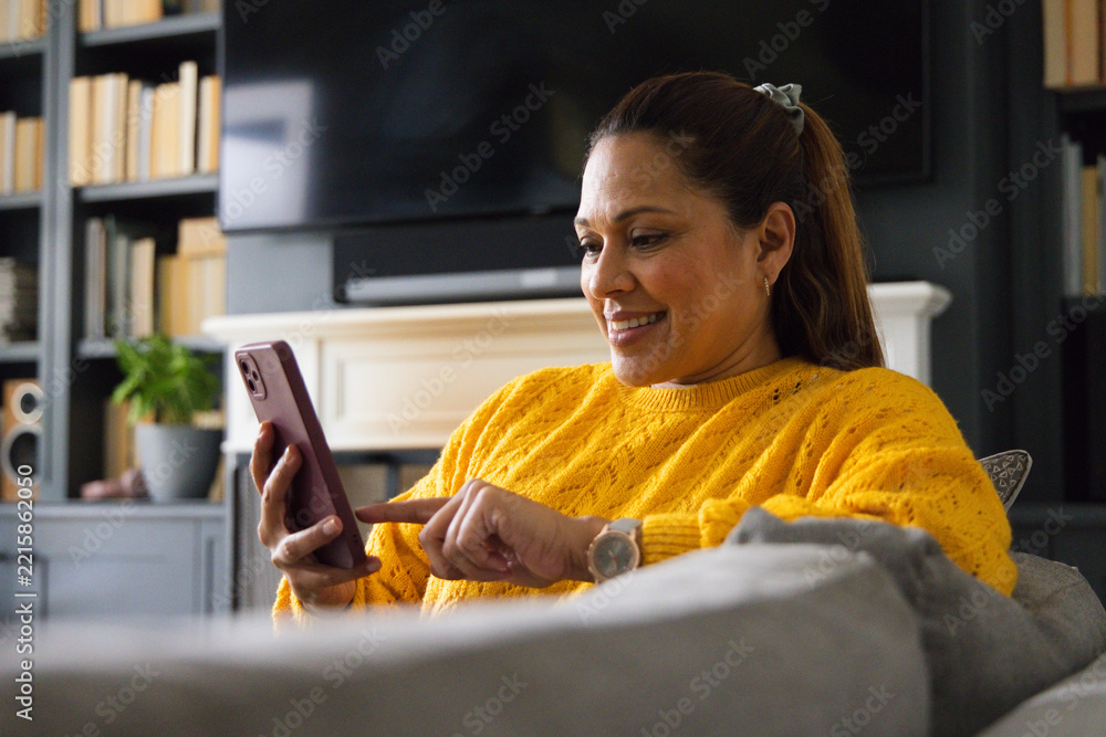 Fototapeta premium Mature African woman sitting on gray couch at home wearing yellow sweater pointing at purple phone