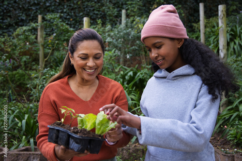 African American mom holding black seed tray while child in pink beanie checking plants in backyard