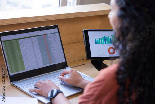 African American woman typing on laptop at desk, viewing tablet charts, rust top, watch, copy space