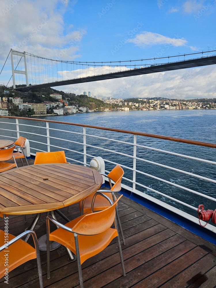 Fototapeta premium Boat deck overlooks bridge and water. Orange chairs surround wooden table. Sky blue with clouds above. Bridge spans distant shoreline in Istanbul, Turkey