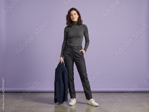 Full length portrait of a stylish young woman in a grey turtleneck and trousers holding a navy blue blazer against a solid purple studio background
