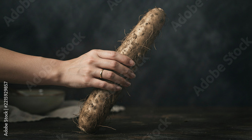 Hand with a gold ring holding a rough root vegetable over dark wood by a linen covered bowl