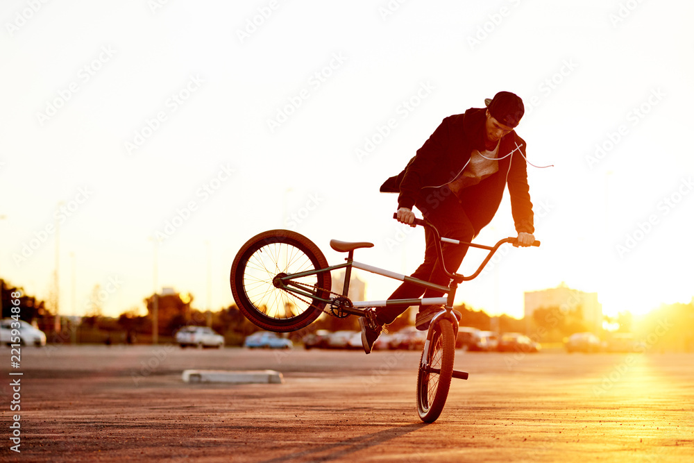 Naklejka premium City, skill and sunset with man on BMX for adrenaline, exercise or extreme sports outdoor. Balance, bike and sunset with rider in parking lot of urban town for action, training or trick practice