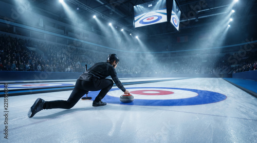 Curler releases stone on ice in competitive sports arena with spectators