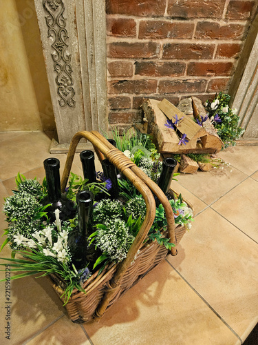 Woven basket holds dark bottles nestled in greenery and flowers. Purple blooms and white blossoms accent the arrangement. Bottles rest among grasses and ferns in a rustic basket