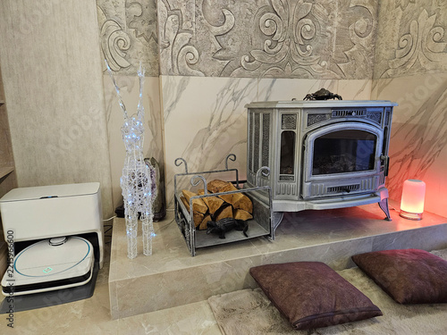 Cozy indoor scene with decorative stove and glowing lantern. Stacked pillows sit on tiled floor beside heater. Shiny wireframe figure stands beside appliance. Firewood rests in metal rack nearby