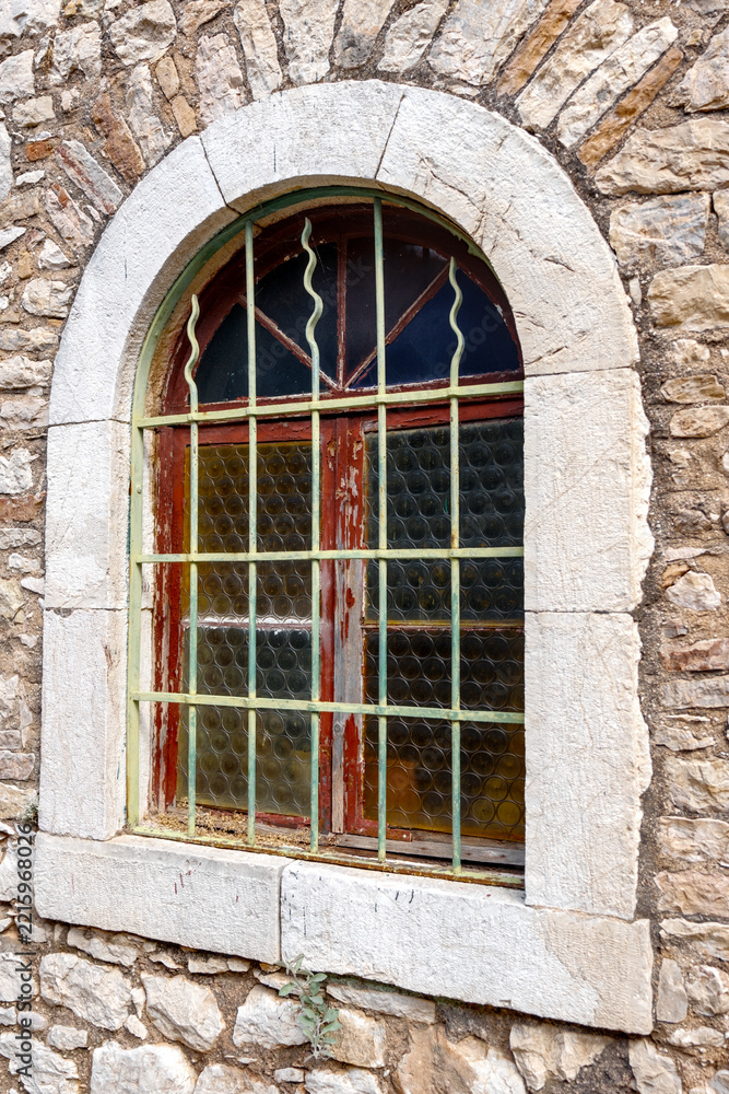 Obraz premium Arched window with iron grille set in weathered wooden frame within rough natural stone masonry wall in Patras, Greece.