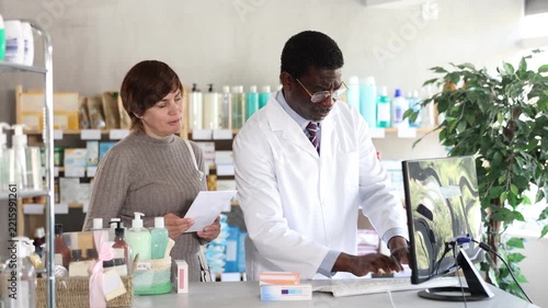 African man pharmacist uses computer at workplace and consulting woman client. Staff reads instructions on computer, interprets medical prescription and makes list of drugs to sell to client 
