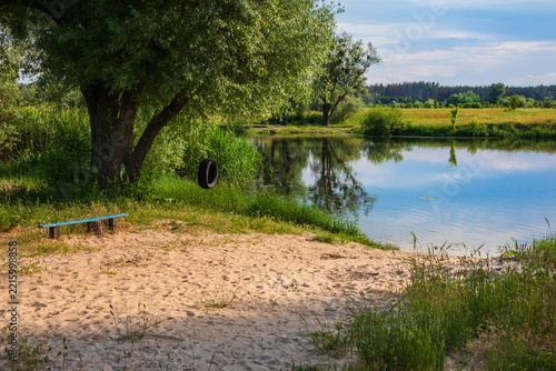 A small, undeveloped riverside beach featuring sand by the water, trees, and green grass. Rural scenery