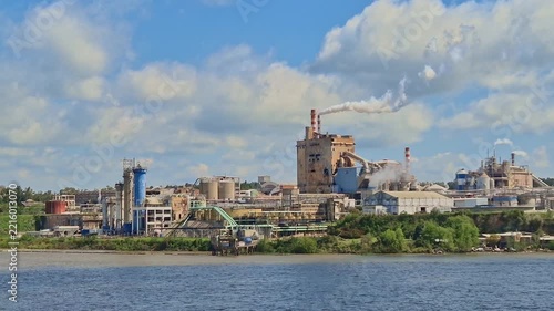 Large old industrial refinery and chemical plant on a riverbank in Argentina, with smoking chimneys, heavy factory structures, and blue sky with clouds
