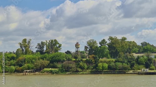 Scenic view of two old vintage water towers rising above lush green trees on a riverbank in Argentina under a cloudy blue sky, captured from water