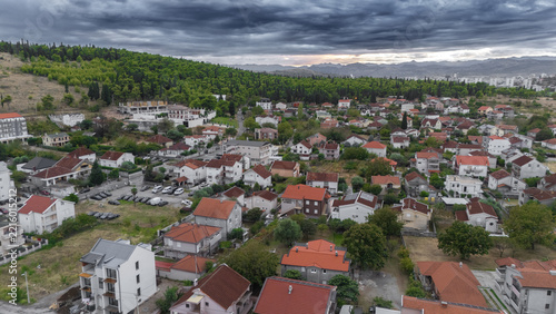 Aerial view of the Zagoric neighborhood featuring residential houses with red-tiled roofs nestled near lush green hills under a dramatic cloudy sky in Podgorica, Tuzi Municipality, Montenegro.