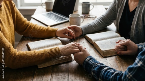 Two people holding hands and praying together over open bibles at a wooden office desk