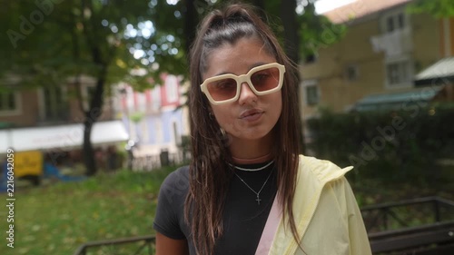 Young woman in cream sunglasses adjusts her look in sunny courtyard portrait with soft garden background