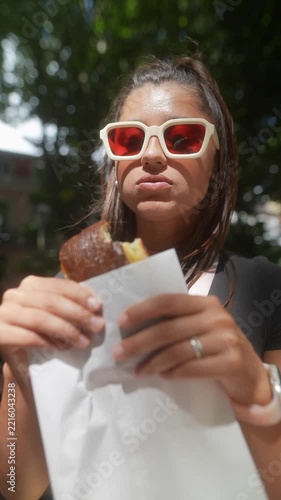 Young woman outdoors in oversized white sunglasses with red lenses holds wrapped pastry near face