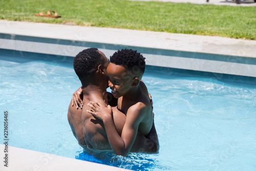African American father and son standing shirtless in backyard pool holding smiling