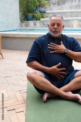 Senior African American man sitting on green yoga mat beside pool wearing navy T-shirt and watch