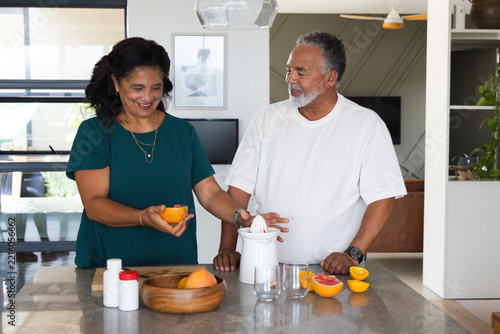 Senior couple preparing fresh citrus juice at kitchen island using white juicer with halved oranges