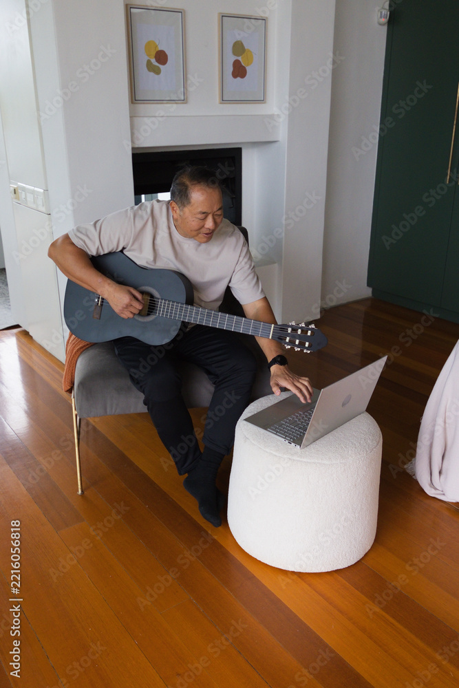 Naklejka premium Senior Asian Chinese man sitting holding blue-gray acoustic guitar and using laptop at home