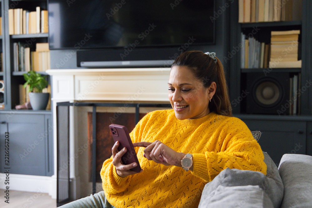 Naklejka premium Mature African American woman in yellow sweater sitting at home on gray sofa tapping purple phone