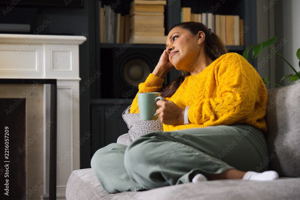 Naklejka premium African American woman sitting on sofa in living room holding mug, in mustard sweater, copy space