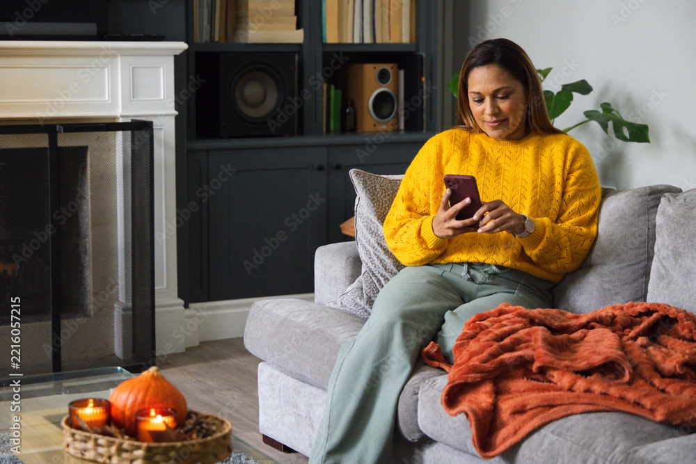 Fototapeta premium African American woman sitting right on sofa wearing mustard yellow sweater using smartphone