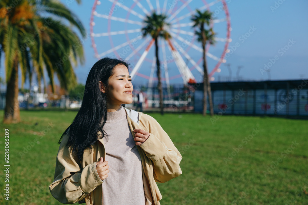 Naklejka premium Woman with backpack enjoying a sunny park day, a ferris wheel in the background, casual jacket, breeze in hair, relaxed mood, travel vibe, outdoor leisure and exploration