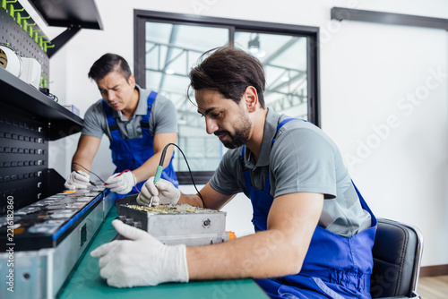 Two technicians in blue overalls solder electronic components at a workstation in a modern factory, focusing on precision assembly and quality control.